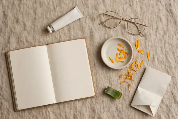 Unguentă fără marcă, notiță goală, ochelari, farfurie ceramică, segment de aloe și petale de calendula pe un prosop de in natural Unguentă fără marcă, notiță goală, ochelari, farfurie ceramică, segment de aloe și petale de calendula pe un prosop de in natural