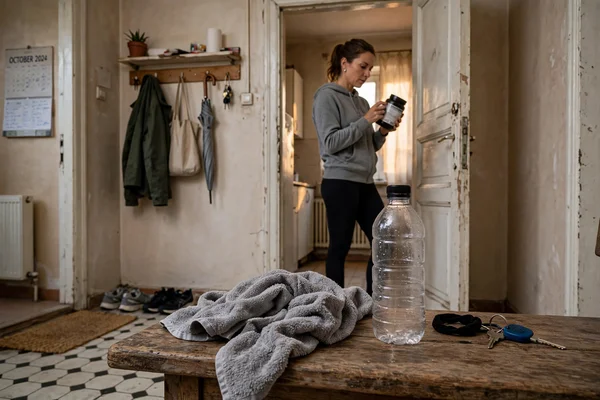 Scenă editorială într-un apartament simplu: un adult activ citește eticheta unui supliment după antrenament, cu o sticlă de apă și un prosop pe o bancă în prim-plan.